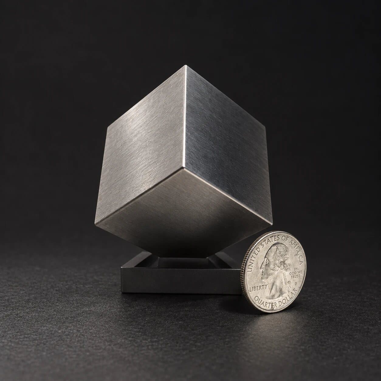 A 1.5 inch brushed tungsten cube balanced on its corner beside a US quarter for scale