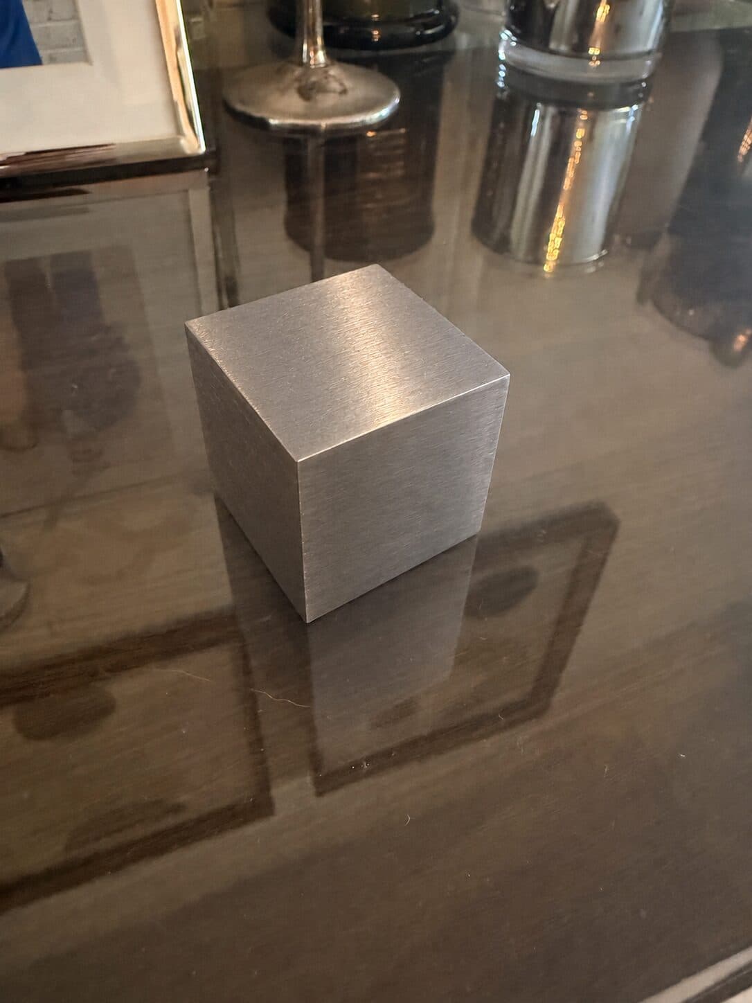 A tungsten cube on a dark glass table, its reflection sharp in the surface