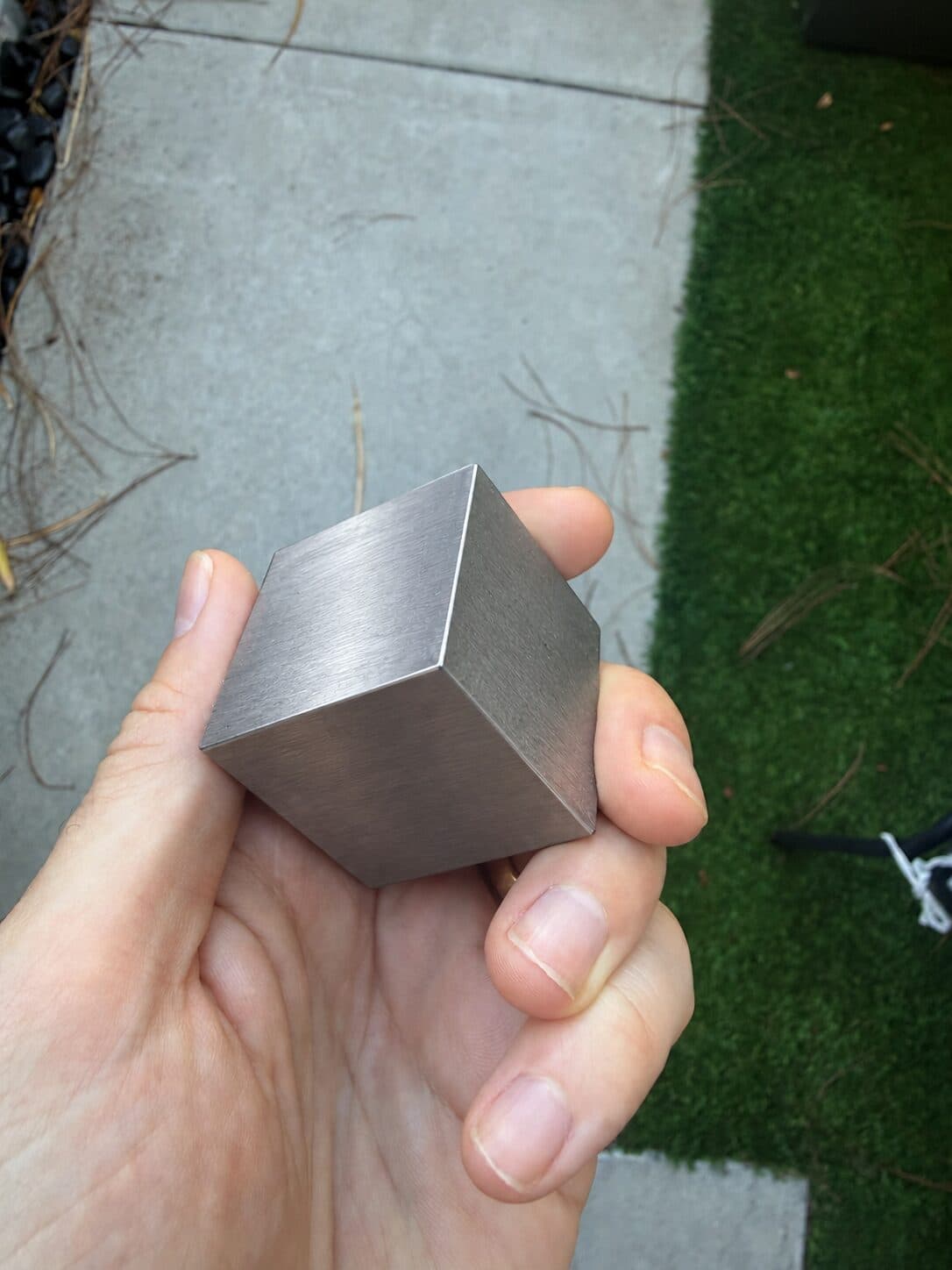 A tungsten cube gripped between thumb and fingers beside a garden path