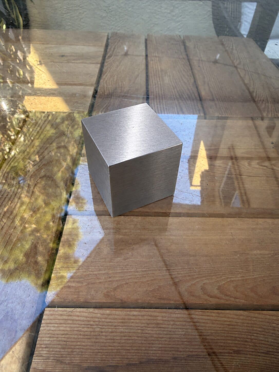 A tungsten cube on a glass table top over a wooden deck in sunlight